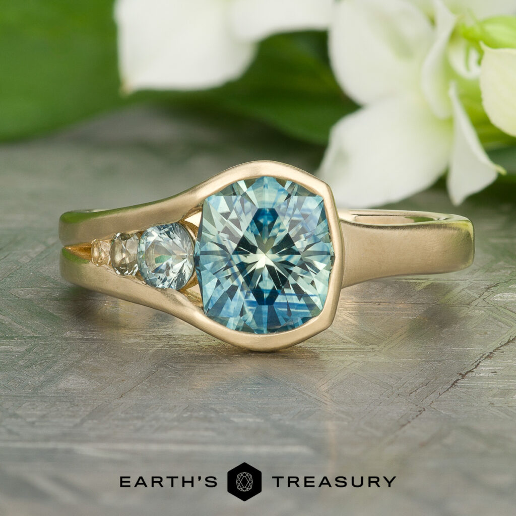 A gold ring with a large blue gemstone at the center, two smaller stones beside it, white flowers in the background, and "Stacked from 26 images. Method=B (R=14,S=2)" displayed below.