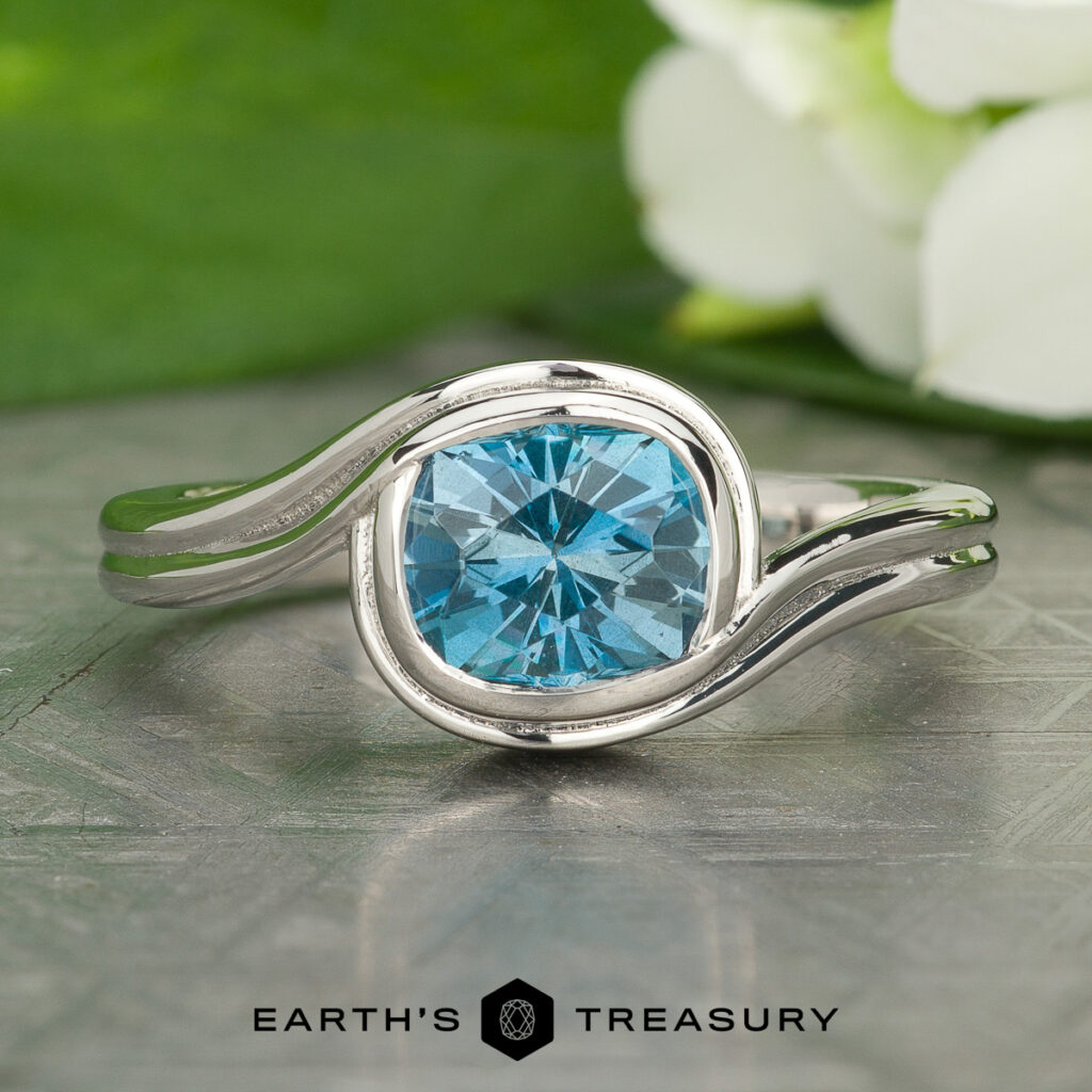 The Stacked from 15 images. Method=B (R=14,S=2) ring features a round blue gemstone set in silver, displayed on a textured surface with green leaves and white flowers in the background.