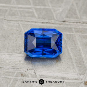 A 1.05-carat, step-cut rectangular blue Ceylon sapphire (heated) is displayed on a textured gray surface, with "EARTH'S TREASURY" at the bottom.