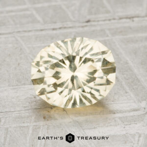 A 1.98-carat faceted light yellow oval Montana sapphire is displayed on a textured light gray surface with "EARTH'S TREASURY" at the bottom.