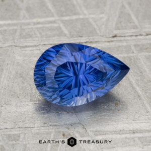 A pear-shaped, vividly blue 2.04-carat Ceylon sapphire rests on a textured metallic surface with "EARTH'S TREASURY" at the bottom.