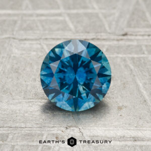 A round, blue 1.10-carat Montana Sapphire (Heated) rests on a textured, light-colored surface with "EARTH'S TREASURY" at the bottom.