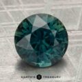 4.29-carat heated sapphire with round cut in blue-green, displayed on a textured gray surface, featuring the "Earth's Treasury" logo and text at the bottom.
