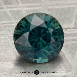 4.29-carat heated sapphire with round cut in blue-green, displayed on a textured gray surface, featuring the "Earth's Treasury" logo and text at the bottom.