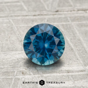 A 1.07-carat heated Montana sapphire with a round brilliant cut is displayed on a textured gray surface, with "EARTH'S TREASURY" at the bottom.