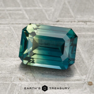 A 2.47-carat heated sapphire with an emerald cut in teal and green rests on a textured surface beside the Earth’s Treasury logo.