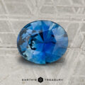 A 2.08-carat round, faceted blue sapphire is displayed on a textured gray surface with the "Earth's Treasury" logo at the bottom.