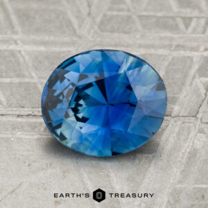 A 2.08-carat round, faceted blue sapphire is displayed on a textured gray surface with the "Earth's Treasury" logo at the bottom.