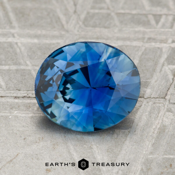 2.08-Carat Sapphire A 2.08-carat round, faceted blue sapphire is displayed on a textured gray surface with the "Earth's Treasury" logo at the bottom.