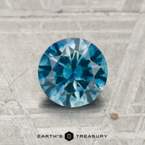 A 1.07-carat heated Montana sapphire in a round brilliant cut is showcased on a textured gray surface with "EARTH'S TREASURY" displayed at the bottom.