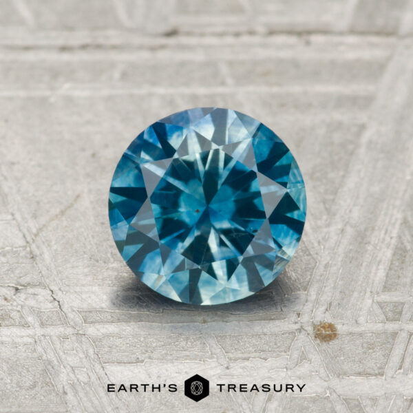 1.07-Carat Montana Sapphire (Heated) A 1.07-carat heated Montana sapphire in a round brilliant cut is showcased on a textured gray surface with "EARTH'S TREASURY" displayed at the bottom.