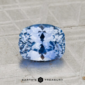 A 2.20-carat Ceylon sapphire with a faceted light blue hue is displayed on a textured, light surface, showcasing the words "EARTH'S TREASURY" at the bottom.