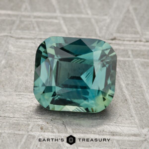 A 2.11-carat heated sapphire with a square cut and blue-green hue rests on a textured gray surface, with "Earth's Treasury" displayed at the bottom.