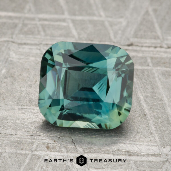 2.11-Carat Sapphire (Heated) A 2.11-carat heated sapphire with a square cut and blue-green hue rests on a textured gray surface, with "Earth's Treasury" displayed at the bottom.