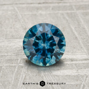 A 1.06-carat heated Montana Sapphire in a round brilliant cut is showcased on a textured surface with the "Earth's Treasury" logo at the bottom.