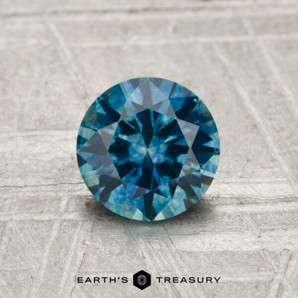 1.06-Carat Montana Sapphire (Heated) A 1.06-carat heated Montana Sapphire in a round brilliant cut is showcased on a textured surface with the "Earth's Treasury" logo at the bottom.
