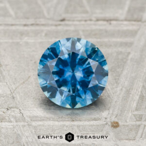 1.05-carat heated Montana sapphire, round brilliant-cut and blue, displayed on a textured light gray surface with the text "Earth's Treasury" below.
