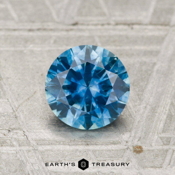 1.05-Carat Montana Sapphire (Heated) 1.05-carat heated Montana sapphire, round brilliant-cut and blue, displayed on a textured light gray surface with the text "Earth's Treasury" below.