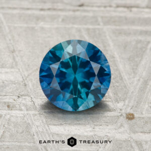 A 1.05-carat heated Montana sapphire, cut in a round brilliant style, is shown on a textured metallic surface with "Earth's Treasury" at the bottom.