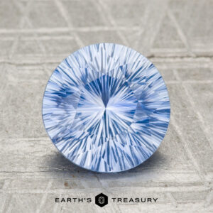 A round, blue-and-white faceted 2.73-carat heated Ceylon sapphire with a starburst pattern rests on a textured light gray surface. "EARTH'S TREASURY" appears below.
