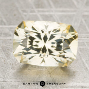 A 4.87-carat Ceylon sapphire, cut in a rectangular faceted shape, rests on a textured gray surface with "Earth's Treasury" written below.