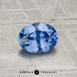 A 1.04-carat Ceylon Sapphire, faceted and oval-shaped, is displayed on a textured light gray surface with the "EARTH'S TREASURY" logo at the bottom.