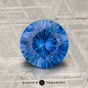 A 1.70-carat heated Ceylon sapphire, round and brilliant-cut, is showcased on a textured silvery surface with "Earth's Treasury" displayed at the bottom.