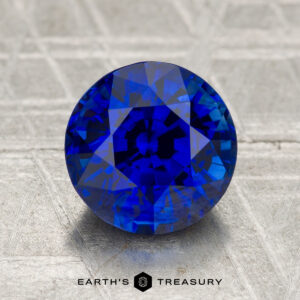 A 2.77-carat heated Ceylon sapphire, round brilliant-cut, sits on a textured metallic surface with "EARTH'S TREASURY" visible below.