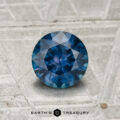 A 1.04-carat heated Montana sapphire with a round cut, shown on a textured gray surface, features "Earth's Treasury" at the bottom.