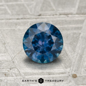 A 1.04-carat heated Montana sapphire with a round cut, shown on a textured gray surface, features "Earth's Treasury" at the bottom.