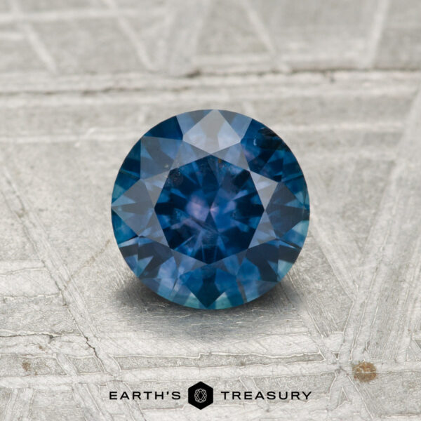 1.04-Carat Montana Sapphire (Heated) A 1.04-carat heated Montana sapphire with a round cut, shown on a textured gray surface, features "Earth's Treasury" at the bottom.