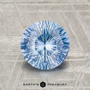 A 1.72-carat heated Ceylon sapphire, round and faceted with a radiant pattern, rests on a textured surface above the "EARTH'S TREASURY" logo and text.