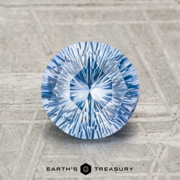 1.72-Carat Ceylon Sapphire (Heated) A 1.72-carat heated Ceylon sapphire, round and faceted with a radiant pattern, rests on a textured surface above the "EARTH'S TREASURY" logo and text.