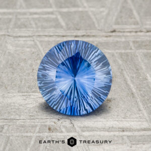 A 1.02-carat Ceylon sapphire (heated), round and faceted, is displayed on a textured gray surface with "EARTH'S TREASURY" at the bottom.