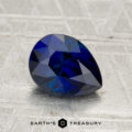 A 1.96-carat Madagascar sapphire (heated), pear-shaped and blue, rests on a textured gray surface with the text "EARTH'S TREASURY" below.