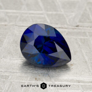 A 1.96-carat Madagascar sapphire (heated), pear-shaped and blue, rests on a textured gray surface with the text "EARTH'S TREASURY" below.