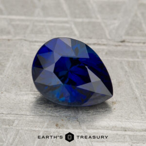 A 1.96-carat Madagascar sapphire (heated), pear-shaped and blue, rests on a textured gray surface with the text "EARTH'S TREASURY" below.