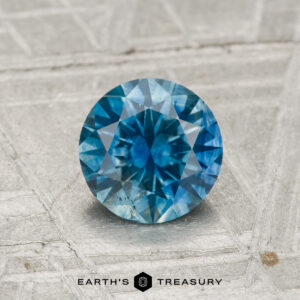 A round brilliant-cut 1.03-carat heated Montana sapphire with a vivid blue hue is displayed on a textured grey surface, featuring the text "Earth's Treasury" below.