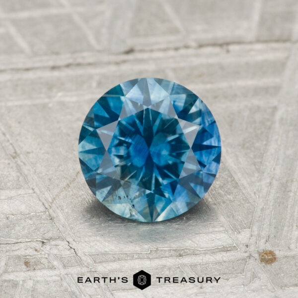 1.03-Carat Montana Sapphire (Heated) A round brilliant-cut 1.03-carat heated Montana sapphire with a vivid blue hue is displayed on a textured grey surface, featuring the text "Earth's Treasury" below.