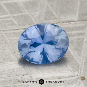 A 1.67-carat Ceylon sapphire, cut in an oval shape, rests on a textured gray surface with "Earth's Treasury" displayed at the bottom.