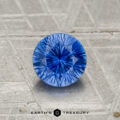 A 1.05-carat heated Ceylon sapphire, round brilliant-cut, is displayed on a textured gray surface with "Earth's Treasury" branding at the bottom.