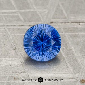 A 1.05-carat heated Ceylon sapphire, round brilliant-cut, is displayed on a textured gray surface with "Earth's Treasury" branding at the bottom.