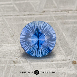 A 1.00-carat Ceylon Sapphire, round brilliant-cut with intricate faceting, rests on a textured gray surface. The "EARTH'S TREASURY" logo appears at the bottom.