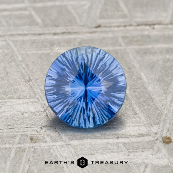 1.00-Carat Ceylon Sapphire A 1.00-carat Ceylon Sapphire, round brilliant-cut with intricate faceting, rests on a textured gray surface. The "EARTH'S TREASURY" logo appears at the bottom.