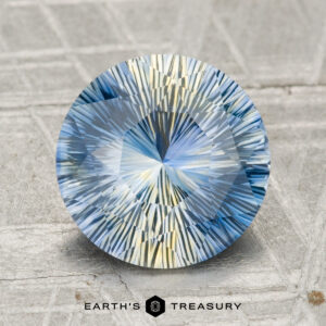 Round, intricately faceted 3.13-carat heated Madagascar sapphire in blue and yellow hues, displayed on a textured surface with the "EARTH'S TREASURY" logo at the bottom.