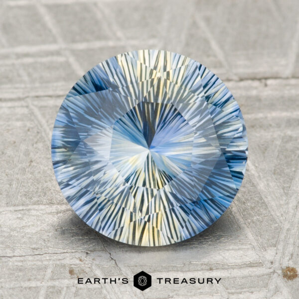 3.13-Carat Madagascar Sapphire (Heated) Round, intricately faceted 3.13-carat heated Madagascar sapphire in blue and yellow hues, displayed on a textured surface with the "EARTH'S TREASURY" logo at the bottom.