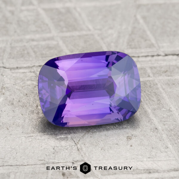 1.51-Carat Ceylon Sapphire A 1.51-carat Ceylon sapphire, faceted and rectangular in shape with a rich purple hue, rests on a textured gray surface. “Earth’s Treasury” appears at the bottom.