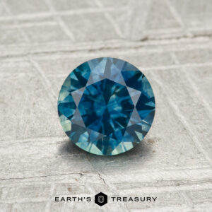A 1.03-carat heated Montana sapphire with a round cut in blue-green, displayed on a textured gray surface with "Earth's Treasury" at the bottom.