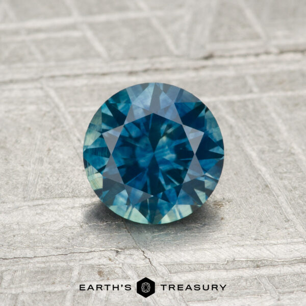 1.03-Carat Montana Sapphire (Heated) A 1.03-carat heated Montana sapphire with a round cut in blue-green, displayed on a textured gray surface with "Earth's Treasury" at the bottom.