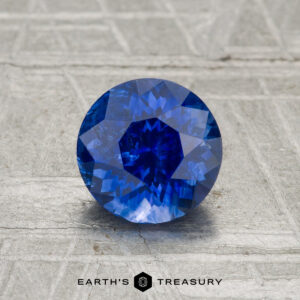 A 1.34-carat heated Ceylon sapphire is displayed on a textured gray surface, with "EARTH'S TREASURY" written below it.